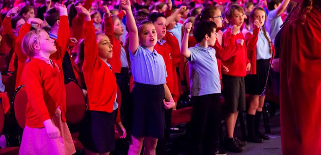 WNO Schools Concert Llandudno Venue Cymru May 21 2018 credit Paul Sampson 48 Paul Sampson 2018 A large group of Primary School children in red and blue uniforms sing, some with arms in the air. Black box rehearsal room setting.