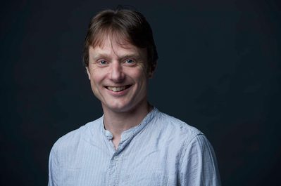 David Adams Head shot of David Adams on a grey background, smiling, wearing a white shirt.