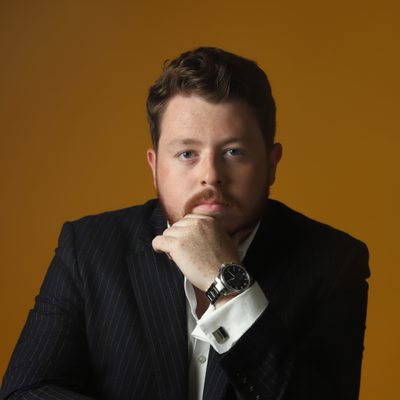 Ross Fettes Headshot A headshot of Ross Fettes, who looks straight at the camera with a serious expression on his face. He wears a smart shirt with cufflinks and a pinstripe suit jacket, and sits on a backwards-facing chair. He leans on this chair with one elbow, his hand supporting his chin.
