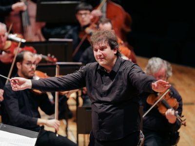 Gi J WNO Orchestra 117 The conductor, Tomas Hanus, side on, holds his arms out wide. There are violin players in the background.