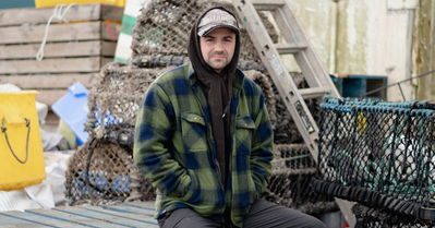 Fisherman's Mission Charity Image A man dressed in a green plaid shirt sits on a boat. Behind him a collection of nets and ladders.