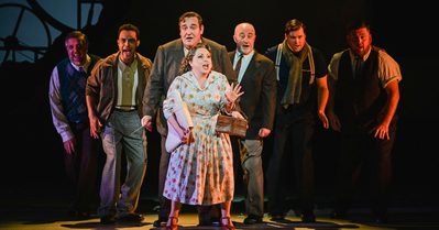 Blaze of Glory Prod Photo A photo from our 2023 production of Blaze of Glory!. Miss Nerys Price stands front and centre, looking excited as she sings. Behind her, Mr Dafydd Pugh and a group of other men also sing passionately.