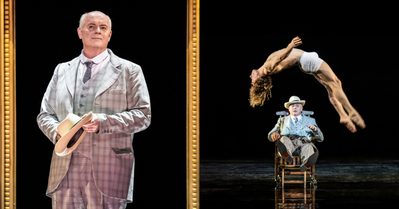 Death In Venice Photos Two photos from Death in Venice. On the left, Gustav von Aschenbach stands, wearing a grey chequered suit and tie and holding his hat. Around him is a large gold frame. On the right, he sits upon a wooden chair, wearing the same suit and hat, as Tadzio, who wears nothing except small white shorts, backflips in front of him.