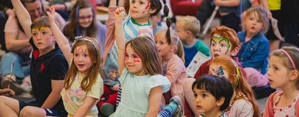 POL Blog 2 Photo 4 A group of young children, some with painted faces, sit together on the floor at Play Opera LIVE. They eagerly watch something that is happening behind the camera, and many have one hand raised in the air, hoping they will be chosen.