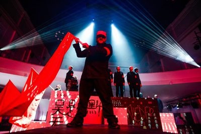 David Pountney Opera Blog An image of multiple men in black standing on a block with Chinese symbols. A man standing in front wave a red flag.
