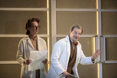 WNO Orlando Sarah Pope WNO Chorus and Daniel Grice Zoroastro Photo Credit Bill Cooper 747 Richard Hubert-Smith 2018 Man in white doctors coat gestrues raising his eyebrows whilst woman with paper stands behind him.