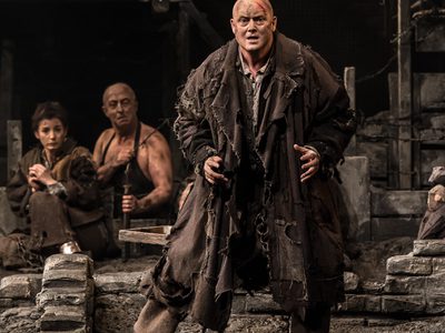 WNO From The House Of The Dead Mark Le Brocq Luka Kuzmich Photo Credit Clive Barda 0101 Clive Barda A chained man, dressed in ragged clothes, looks out to the audience with a pained expression.