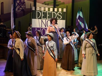WNO Cast Of Rhondda Rips It Up! Photo Credit © Jane Hobson 2524 Jane Hobson 2018 The women's chorus are in traditional dress, marching in front of a green, white and purple 'Deeds Not Words' banner. They are waving green, white and purple flags and wearing Vote for Women Sashes