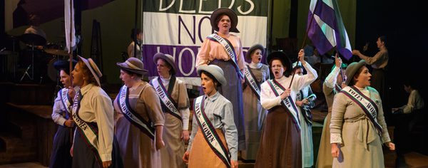 WNO Cast Of Rhondda Rips It Up! Photo Credit © Jane Hobson 2524 Jane Hobson 2018 The women's chorus are in traditional dress, marching in front of a green, white and purple 'Deeds Not Words' banner. They are waving green, white and purple flags and wearing Vote for Women Sashes