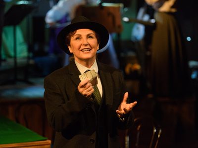 WNO Rhondda Rips It Up! Lesley Garrett Emcee Photo Credit © Jane Hobson 7336 Jane Hobson 2018 Emcee dressed in a dark blazer, bowler hat and green tie, hands raised in front of her holding a letter as she sings to to audience, looking slighting upwards.