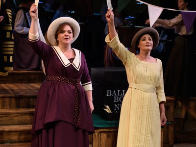 WNO Rhondda Rips It Up! Madeleine Shaw Lady Rhondda And Anitra Blaxhall Helen Archdale Photo Credit © Jane Hobson 7474 Jane Hobson 2018 Lady Rhondda wears a purple dress with white collar and white hat , Helen Archdale wears a yellow, long sleeved dress and brown hat. They stand in front of the polling station, both holding voting cards in the air.
