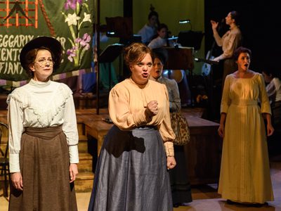WNO Rhondda Rips It Up! Rosie Hay Sybil Madeleine Shaw Lady Rhondda Catherine Wood Lottie And Anitra Blaxhall Helen Archdale Photo Credit © Jane Hobson 2730 Jane Hobson 2018 Sybil is dressed in a white frilled blouse and brown skirt, Lady Rhondda is dressed in a beige blouse and blue skirt. They look out to the distance. Lottie and Helen Archdale stand slightly behind in the darker light watching on.