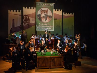 WNO Rhondda Rips It Up! WNO Ladies Chorus Photo Credit © Jane Hobson 2700 Jane Hobson 2018 The image shoes a backdrop of a 'Cat and Mouse Poster' with the orchestra in the background.The ladies chorus are sat around a billiard table dressed in suits. They gather around holding cardboard mice with one holding a cardboard cat.