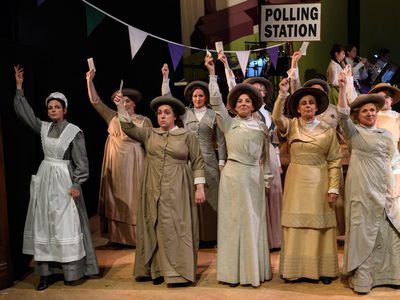 WNO Rhondda Rips It Up! WNO Ladies Chorus Photo Credit © Jane Hobson 2846 Jane Hobson 2018 Women's chorus are stood at the polling station , all holding their voting cards, and facing the audience.
