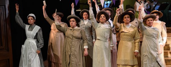 WNO Rhondda Rips It Up! WNO Ladies Chorus Photo Credit © Jane Hobson 2846 Jane Hobson 2018 Women's chorus are stood at the polling station , all holding their voting cards, and facing the audience.