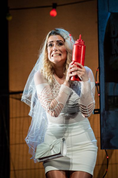 WNO Don Pasquale Harriet Eyley Norina Photo credit Robert Workman 088 Robert Workman 2019 Woman in short white dress and veil holding red ketchup bottle grimacing.