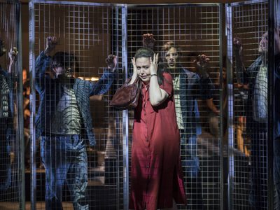 WNO Dead Man Walking Lucia Cervoni Sister Helen Prejean Photo credit Johan Persson 0923 Johan Persson 2019 Woman holds her head between her hands as men lean against metal fence behind.