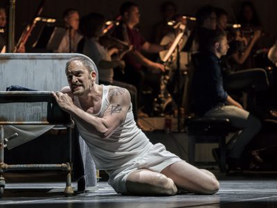 WNO Dead Man Walking Morgan Smith Joseph De Rocher Photo credit Johan Persson 02075 Johan Persson 2019 Man in white vest and shorts sits on the floor grasping onto bed anguished.