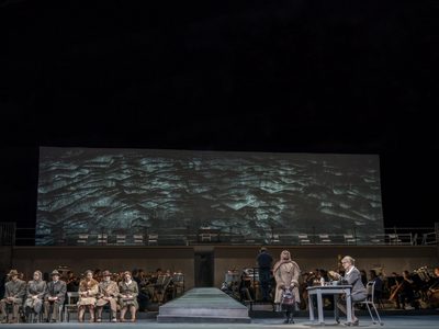 WNO The Consul Cast of The Consul Photo credit Johan Persson 01366 Johan Persson 2019 Wide shot of stage woman standing infront of desk whilst others are seated on the other side.