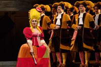 WNO La Cenerentola Heather Lowe Tisbe WNO Chorus Photo credit © Jane Hobson 1760 Jane Hobson Tisbe wears a pink, yellow and orange velvet outfit. She has big, yellow hair and a tiny pink top hat. She is grimacing.