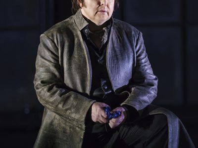 WNO Roberto Devereux Barry Banks Roberto Devereux Photo Credit Bill Cooper 0795 Bill Cooper 2019 Man stood with one foot raised on something in dark leather coat.