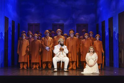 WNO The Magic Flute James Platt Sarastro Anita Watson Pamina Photo credit Bill Cooper 0258 Bill Cooper 2019 Woman in white kneeling at front, man sat in white, chorus standing in orange behind.
