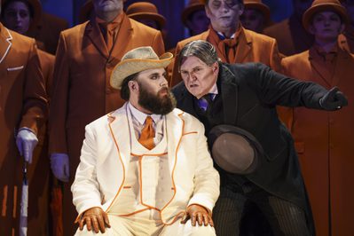 WNO The Magic Flute James Platt Sarastro Howard Kirk Monostatos Photo credit Bill Cooper 0718 Bill Cooper 2019 Man in white outfit is seated whilst man in black suit leans down talking to him pointing.