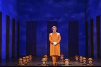 WNO The Magic Flute Simon Cosby Buttle First Priest Chorus Photo credit Bill Cooper 0411 Bill Cooper 2019 Man in orange clothes stands while other heads with orange bowler hats poke through the floorboards.