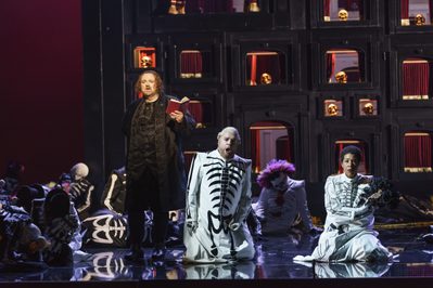 WNO Un ballo in maschera Gwyn Hughes Jones Riccardo Roland Wood Renato Mary Elizabeth Williams Amelia Photo credit Bill Cooper© 0545 Bill Cooper 2019 Skeleton figures kneel on the floor as a man dressed in black stands singing