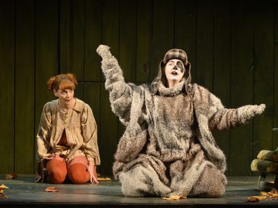 Aoife Miskelly as Vixen Helen Greenway as Dog The Cunning Little Vixen Photocredit Richard Hubert Smith 9262 Richard Hubert-Smith 2019 Dog dressed character kneels arms open front of stage while fox character kneels behind.