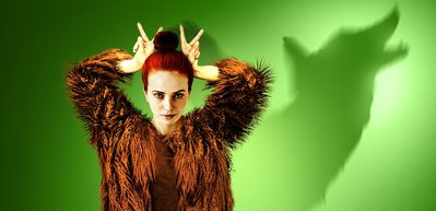 WNO 1000x486 Vixen Aoife Miskelly as Vixen A young woman is dressed in brown fur. In the green background is the silhouette of a wolf.