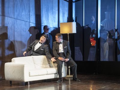 WNO Rigoletto 2019 David Junghoon Kim The Duke and Simon Crosby Buttle Borsa Photocredit Richard Hubert Smith 3351 Richard Hubert-Smith 2019 Dark party scene two men singing over white sofa.