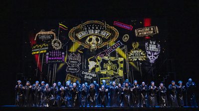 WNO Candide Cast of Candide photo credit Johan Persson 03611 A group in long black coats stand before a display of colourful Spanish and Latin American inspired displays