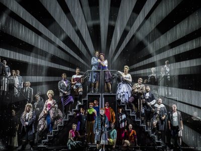 WNO Candide Cast of Candide photo credit Johan Persson 10631 A group of people stand on two sets of stairs, with light radiating from them in a circle