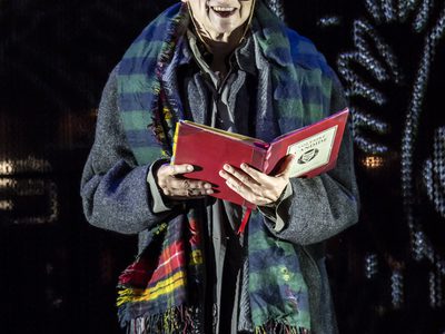 WNO Candide Gillian Bevan Pangloss photo credit Johan Persson 00834 A woman with bright orange hair reads from a red book