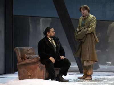 WNO La bohème Benson Wilson Schaunard David Shipley Colline photo credit Richard Hubert Smith 4221 A man in a black coat sits on a brown leather armchair, answering questions in the snow