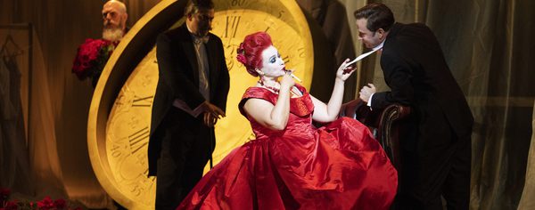 WNO The Makropulos Affair Cast of The Makropulos Affair photo credit Richard Hubert Smith 1775 Woman in red dress pulls a man's tie seductively, in front of a giant clock
