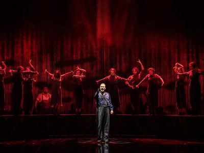 Alfredo Tejada as Ruiz Alonso in WNO Ainadamar 2023 Credit Johan Persson 13495 A man dressed as a Spanish police officer reaches out to the front of stage, the people behind are bathed in red light