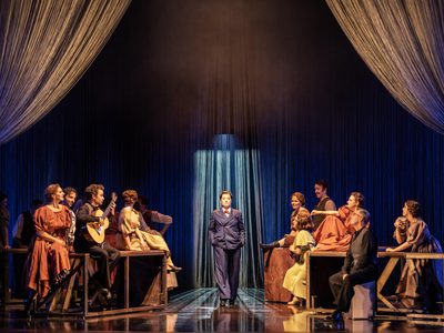 Hanna Hipp as Federico García Lorca in WNO Ainadamar 2023 Credit Johan Persson 06243 a woman in a suit walks forwards in a bar scene, as the patrons observe them
