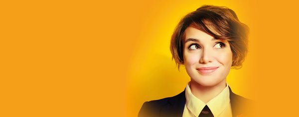 WNO CL1881 S24 Website image 2000x785 Header Così fan tutte A woman from cosi fan tutte wears a suit and tie. She is smirking and is against a yellow background