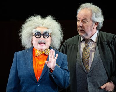 WNO Così fan tutte Rebecca Evans Despina José Fardilha Don Alfonso photo credit Elliott Franks 1967 A production shot of a woman dressed as a man, overlooked by a man in a suit and graduation gown