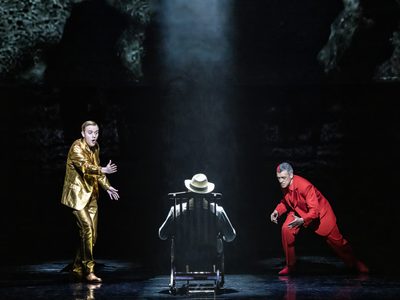 WNO Death in Venice Alexander Chance The Voice of Apollo Mark le Brocq Gustav von Aschenbach Roderick Williams The Voice of Dionysus photo credit Johan Persson 07250 A production shot of three men- one sits in a chair with his back to the audience as the other two gesture to him.