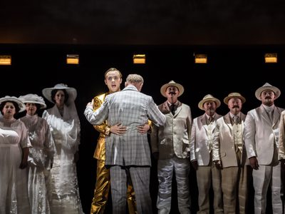 WNO Death in Venice Alexander Chance The Voice of Apollo Mark le Brocq Gustav von Aschenbach and the cast of Death in Venice photo credit Johan Persson 04404 A production shot shows a group of men and women in pale outfits with a man in a gold suit hugging one of them