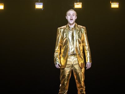 WNO Death in Venice Alexander Chance The Voice of Apollo photo credit Johan Persson 03811 A production shot showing a man in a gold suit singing to the audience
