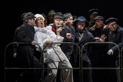 WNO Death in Venice Roderick Williams The Old Man and the Cast of Death in Venice photo credit Johan Persson 01210 A production shot where a chef leans back into a group of men dressed in dark clothing. They al look shocked.