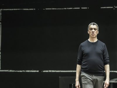 WNO Death in Venice Roderick Williams Traveller photo credit Craig Fuller 9849 A rehearsak shot of Roderick Williams stood with a serious expression in front of a black board