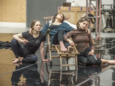 WNO Death in Venice Vilhelmiina Sinervo Daughter 2 Diana Salles The Polish Mother Selma Hellmann Daughter 1 photo credit Craig Fuller0054 A rehearsal shot shows three woman leaning into each ither sat on the floor and a chair.