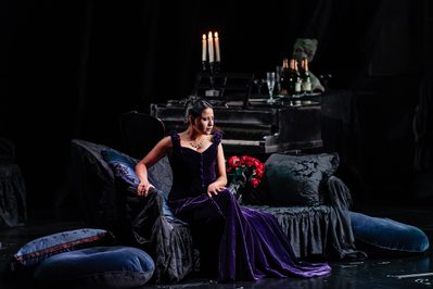 WNO La traviata Stacey Alleaume Violetta photo credit Julian Guidera 7 A woman in a black dress sits upon a dark sofa on a dark background