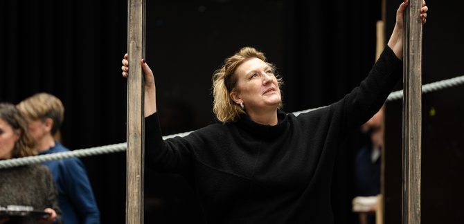 WNO Peter Grimes Dame Sarah Connolly Auntie photo credit Dafydd Owen 1160 Dame Sarah Connolly stands in a doorframe during rehearsals. She holds its sides and gazes upwards.