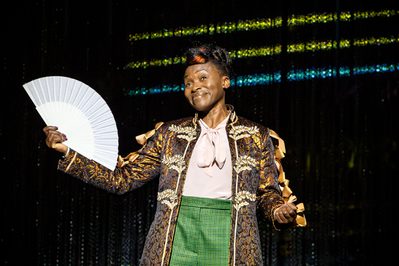 WNO Candide Rakie Ayola Pangloss Narrator Sage Beggar photo credit Craig Fuller 1219 The Narrator/Pangloss smiles contentedly out to the audience, holding a fan in her right hand.